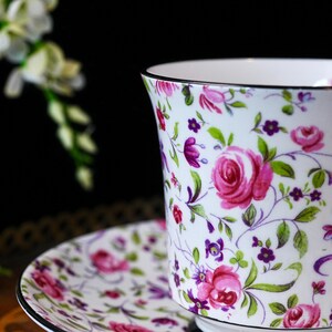 May include: A white teacup and saucer set with a floral design. The pattern includes pink roses, purple flowers, and green leaves on a white base. The cup and saucer have a black trim.
