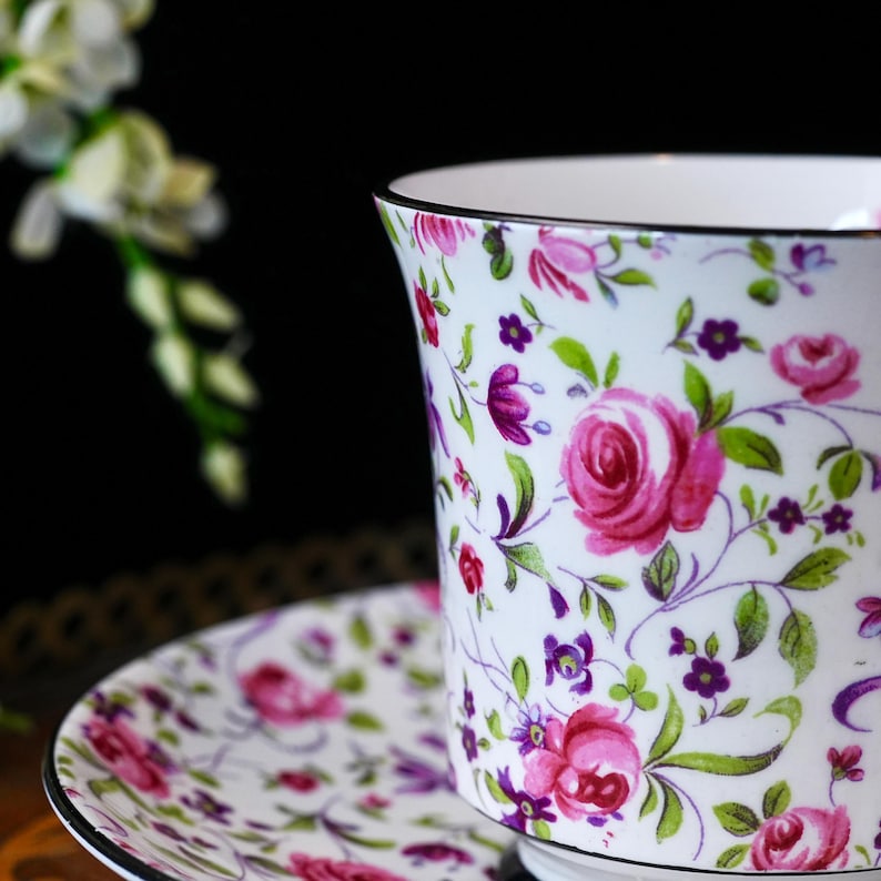 May include: A white teacup and saucer set with a floral design. The pattern includes pink roses, purple flowers, and green leaves on a white base. The cup and saucer have a black trim.