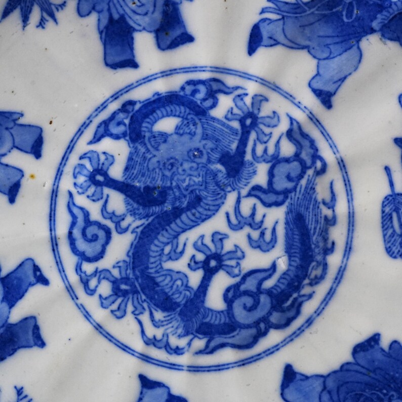 Puede incluir: Un plato de cer&aacute;mica blanco con un dise&ntilde;o de drag&oacute;n azul en el centro. El drag&oacute;n est&aacute; rodeado por un borde circular y patrones florales. El plato tiene un borde festoneado y motivos florales azules adicionales.