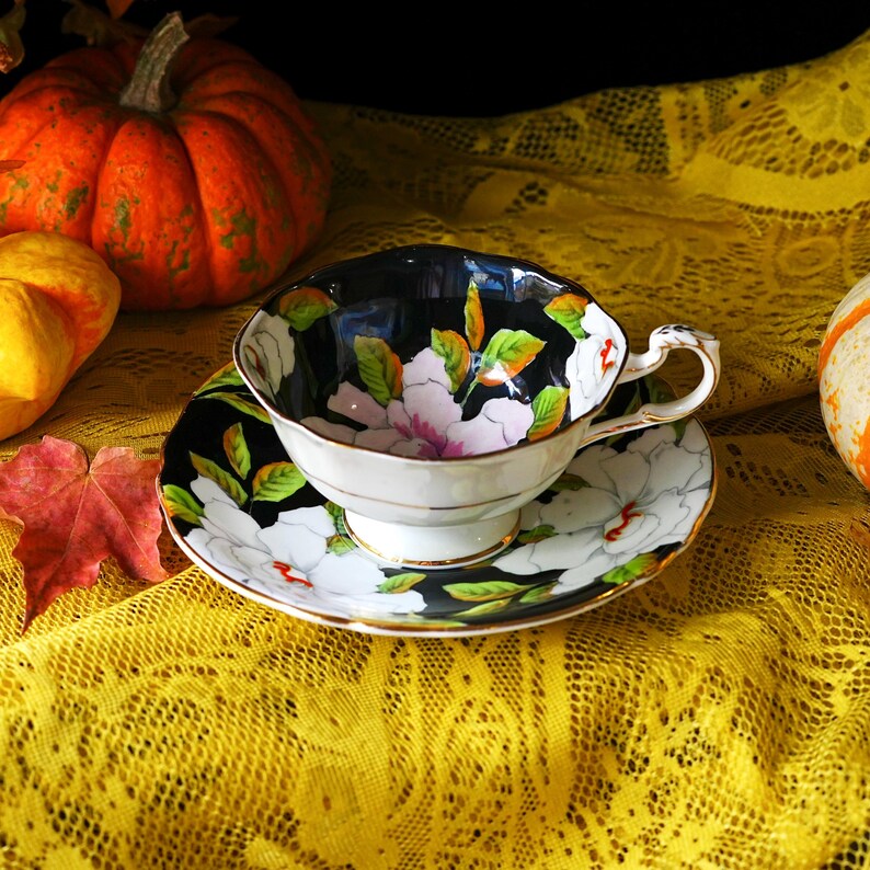 May include: A vintage teacup and saucer set with a black interior and floral design. The cup and saucer feature white flowers with pink centers and green leaves, accented with gold trim. The set is placed on a yellow lace tablecloth, with pumpkins in the background.