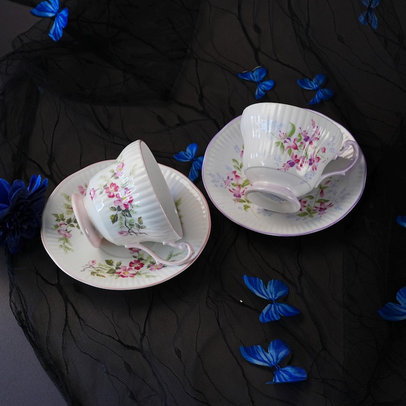 May include: Two white porcelain teacups and matching saucers featuring a ribbed texture and a delicate pink and green floral pattern. Each cup has a curved handle and a light pink base, displayed alongside several small blue butterfly decorations.
