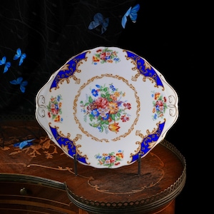 Puede incluir: Una bandeja de servicio de porcelana blanca con borde festoneado, que presenta un ramo central de flores coloridas, un borde ancho azul cobalto y detalles intrincados de filigrana dorada a lo largo del borde y las asas.