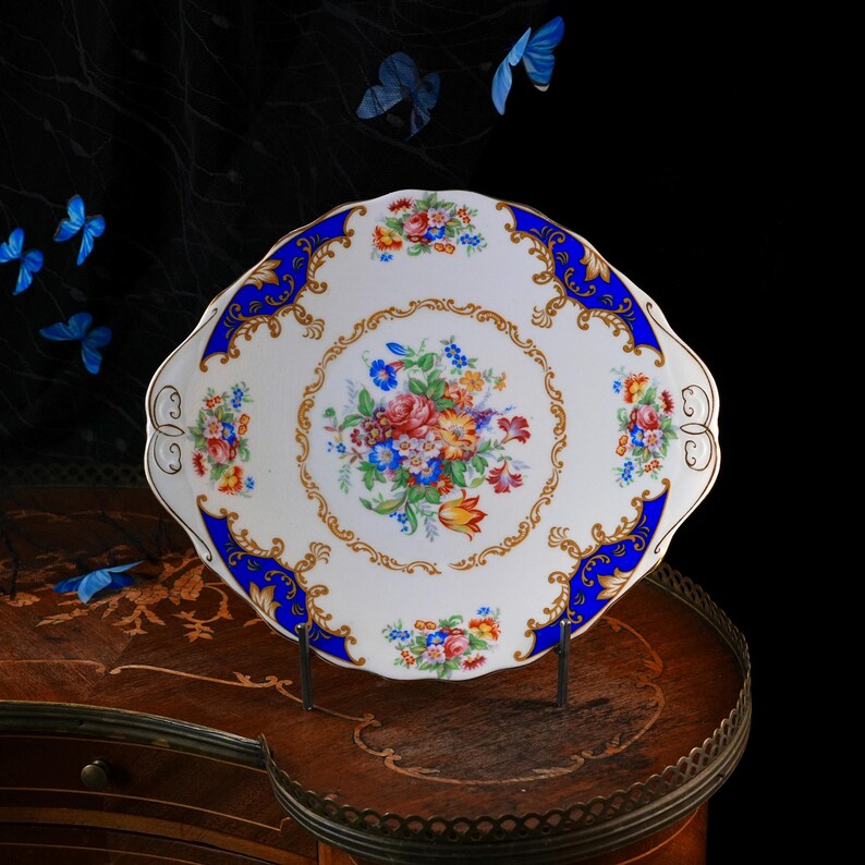 Puede incluir: Una bandeja de servicio de porcelana blanca con borde festoneado, que presenta un ramo central de flores coloridas, un borde ancho azul cobalto y detalles intrincados de filigrana dorada a lo largo del borde y las asas.