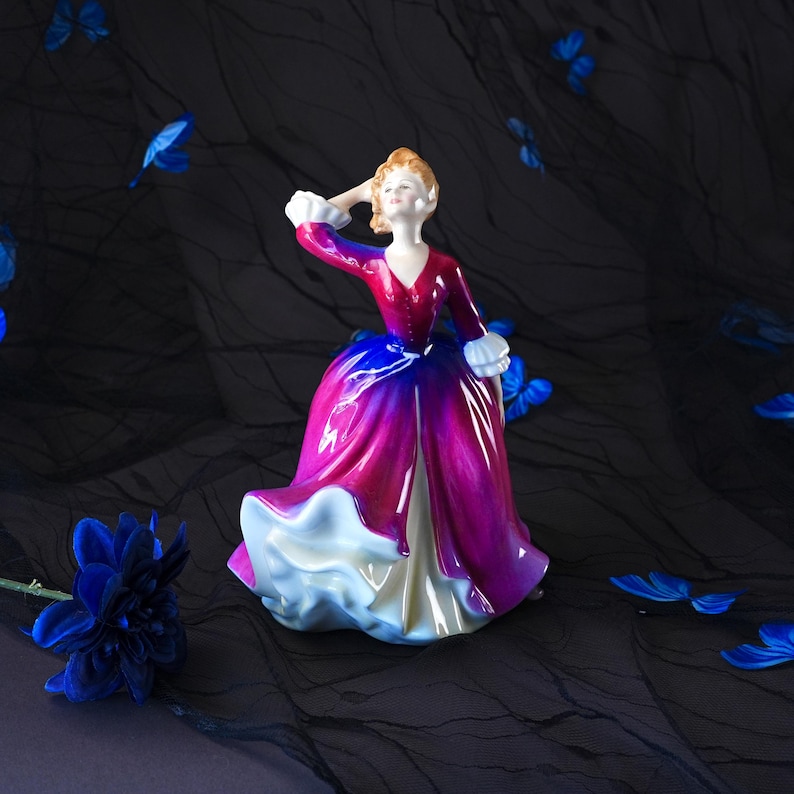 May include: A porcelain figurine of a person with light brown hair wearing a long dress that transitions from a deep red bodice to a vibrant magenta and blue skirt with a white ruffled hem and white cuffs, posed with one hand near their head.