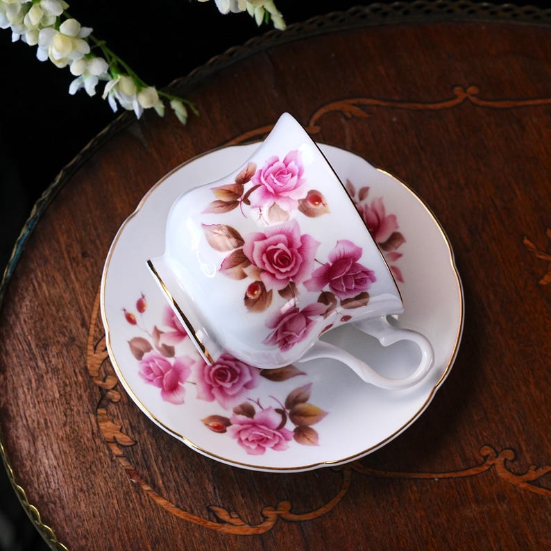 Op de afbeelding: Witte porseleinen theekop en schotel set versierd met roze rozen en gouden rand. De theekop heeft een hartvormig handvat. De set staat op een houten oppervlak.