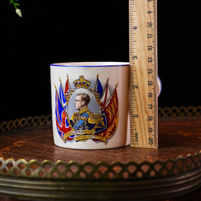 Op de afbeelding: Een witte keramische mok met een portret van koning Edward VIII, omringd door vlaggen en een kroon. De mok heeft een blauwe rand en een oor. De mok is ongeveer 7.6 cm hoog.
