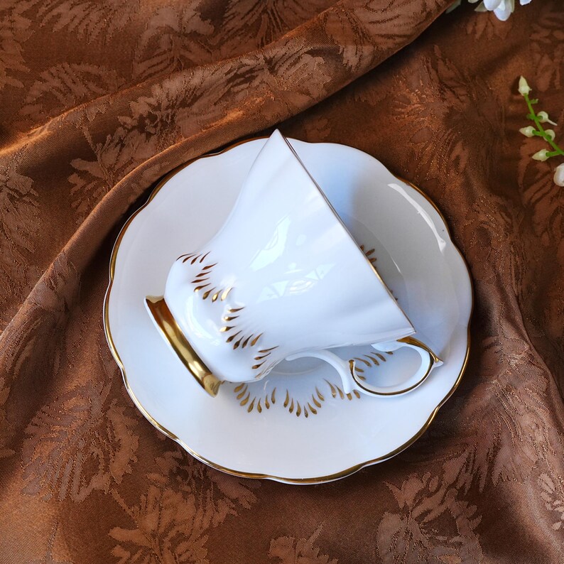 May include: A white teacup and saucer set with gold trim and accents. The teacup is tilted, revealing its interior. The saucer and cup have a scalloped edge. The set is placed on a brown, patterned surface.