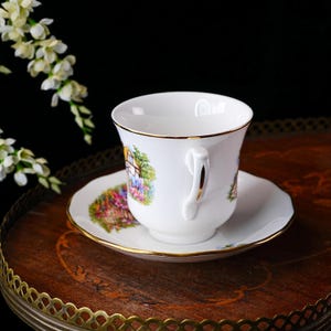 May include: White teacup and saucer set with gold trim. The cup and saucer feature a colorful floral and cottage scene. The set is displayed on a wooden tray with a decorative metal edge. A sprig of white flowers is in the background.