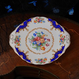Puede incluir: Un plato de servicio redondo de porcelana con borde festoneado y asas integradas. El centro presenta un colorido ramo de flores, rodeado por un borde de filigrana dorada y un borde ancho azul cobalto con detalles dorados y motivos florales.