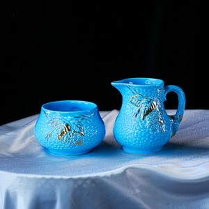Op de afbeelding: Een felblauwe keramische melkkan en een bijpassende suikerpot. Beide stukken hebben een getextureerd, korrelig oppervlak met reli&euml;f en goudkleurige blad- en bessenmotieven. De kan heeft een gebogen handvat en een schenktuit, de pot is rond.