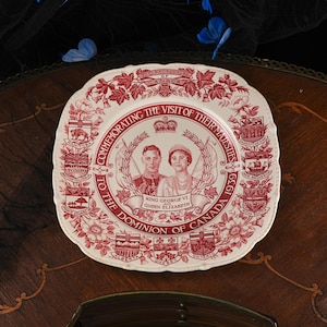 May include: A square ceramic plate with a cream background and deep red transferware print. The centre features portraits of King George VI and Queen Elizabeth, surrounded by text, a crown, and the coats of arms of various Canadian provinces.