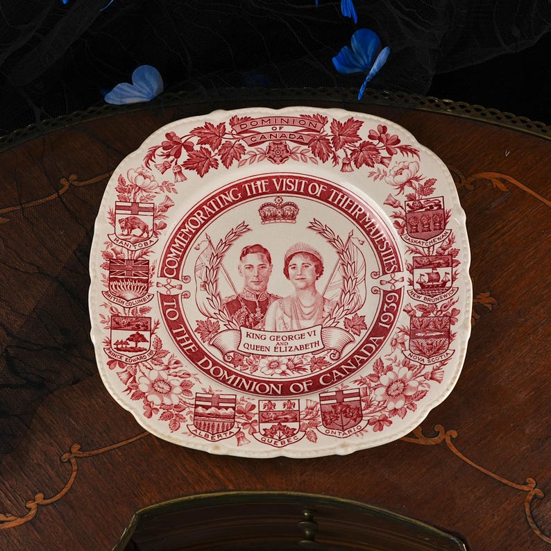 May include: A square ceramic plate with a cream background and deep red transferware print. The centre features portraits of King George VI and Queen Elizabeth, surrounded by text, a crown, and the coats of arms of various Canadian provinces.
