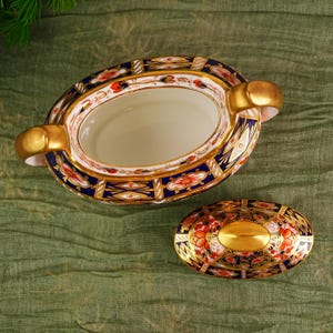 May include: An oval-shaped, antique porcelain serving dish with a matching lid. The dish features a white interior, gold handles, and is decorated with a blue, red, and gold floral pattern. The lid has a gold oval center.