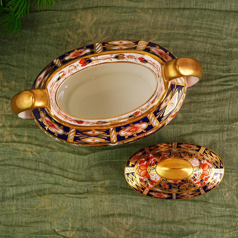May include: An oval-shaped, antique porcelain serving dish with a matching lid. The dish features a white interior, gold handles, and is decorated with a blue, red, and gold floral pattern. The lid has a gold oval center.