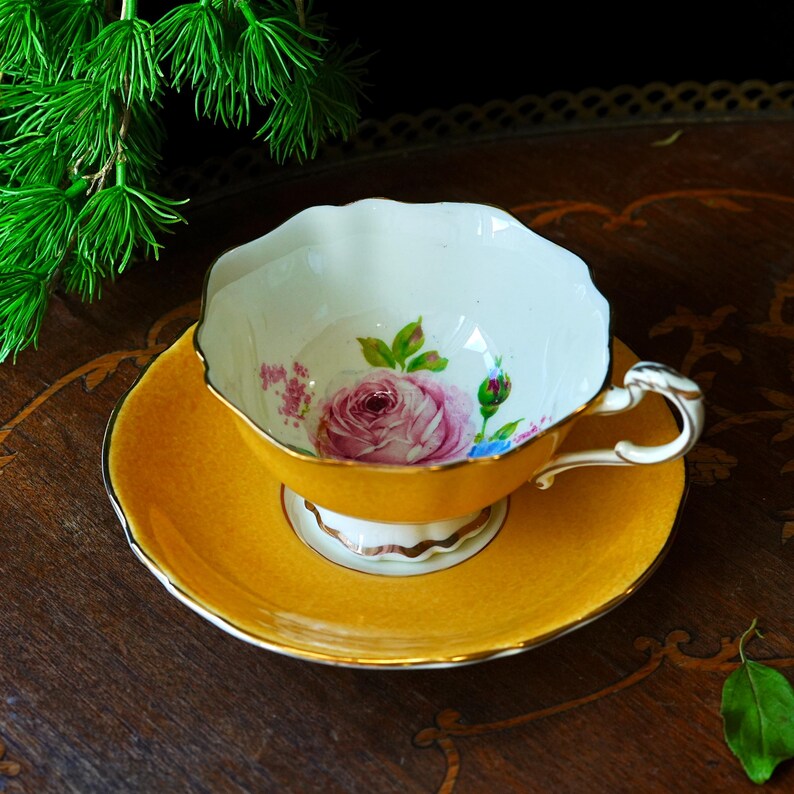 May include: A vintage teacup and saucer set. The cup and saucer are a bright yellow with gold trim. The inside of the cup features a pink rose design. The handle of the cup is white with gold accents. A classic tea set.