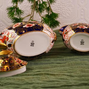 May include: Two ornate porcelain teacups with lids, featuring a vibrant floral pattern in red, gold, and blue. The teacups are set on a green cloth, showcasing their intricate design and craftsmanship. The lid is off one of the teacups.