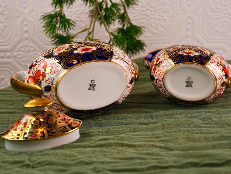 May include: Two ornate porcelain teacups with lids, featuring a vibrant floral pattern in red, gold, and blue. The teacups are set on a green cloth, showcasing their intricate design and craftsmanship. The lid is off one of the teacups.