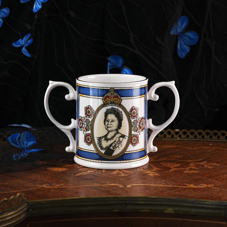 May include: A white porcelain two-handled mug featuring a central oval portrait of Queen Elizabeth the Queen Mother wearing a crown and jewellery. The design includes blue borders, red and gold floral motifs, and a gold crown above the portrait.