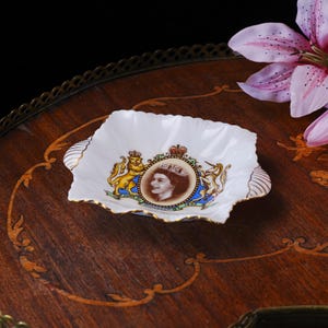 May include: A white porcelain square dish with scalloped edges and gold trim. The center features a portrait of Queen Elizabeth II within an oval frame, flanked by a lion and a unicorn, with the text June 2nd 1953 printed on a blue banner.
