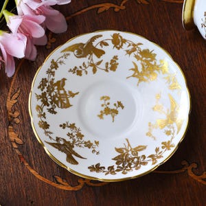Op de afbeelding: Witte schotel met een gouden rand en ingewikkelde gouden vogel- en bloemmotieven. De geschulpte rand voegt een vleugje elegantie toe aan dit decoratieve stuk. Een klassiek ontwerp voor een theeservies.