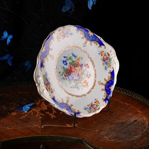 Puede incluir: Un plato de porcelana blanca con borde festoneado, que presenta un ramo floral central de flores rojas, rosas, amarillas y azules. El borde est&aacute; decorado con paneles de color azul intenso, filigranas doradas y motivos florales.