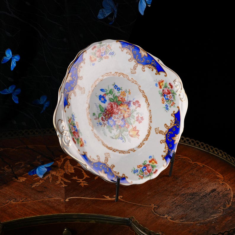 Puede incluir: Un plato de porcelana blanca con borde festoneado, que presenta un ramo floral central de flores rojas, rosas, amarillas y azules. El borde est&aacute; decorado con paneles de color azul intenso, filigranas doradas y motivos florales.