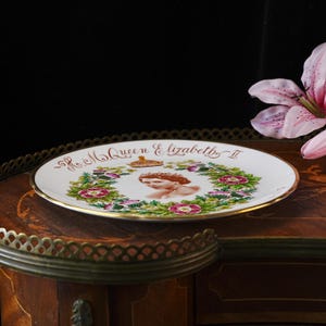 Op de afbeelding: Een wit keramieken herdenkingsbord met een centraal portret van koningin Elizabeth II, een gouden kroon en een bloemenkrans van rozen en distels. De tekst H.M. Queen Elizabeth II staat in gouden letters aan de bovenrand van het bord.