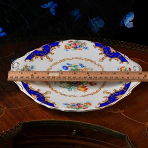 Puede incluir: Un plato de servicio de porcelana blanca con borde festoneado, con un patr&oacute;n floral colorido en el centro y bordes azul cobalto con volutas doradas. Una regla de madera est&aacute; colocada sobre el plato, mostrando un di&aacute;metro de 25,4 cm.