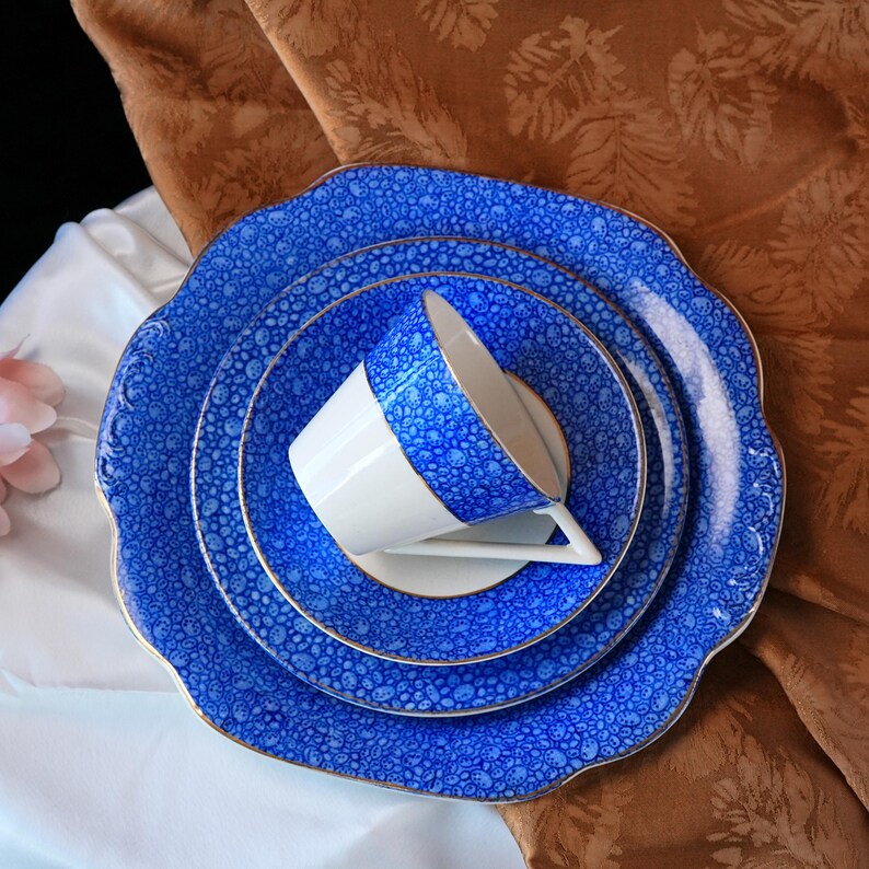 May include: A blue and white porcelain tea set with a floral pattern. The set includes a teacup, saucer, and three plates. The edges of the plates and cup are scalloped and trimmed with gold. The background is a brown and white floral fabric.