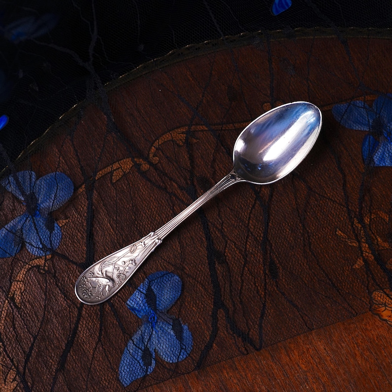 Puede incluir: Una cuchara de metal plateado con un mango alargado que presenta un motivo floral y de ave en relieve. La cuchara tiene un cuenco ovalado liso y reflectante, y un cuello delgado que se ensancha hacia un extremo decorado con detalles intrincados.