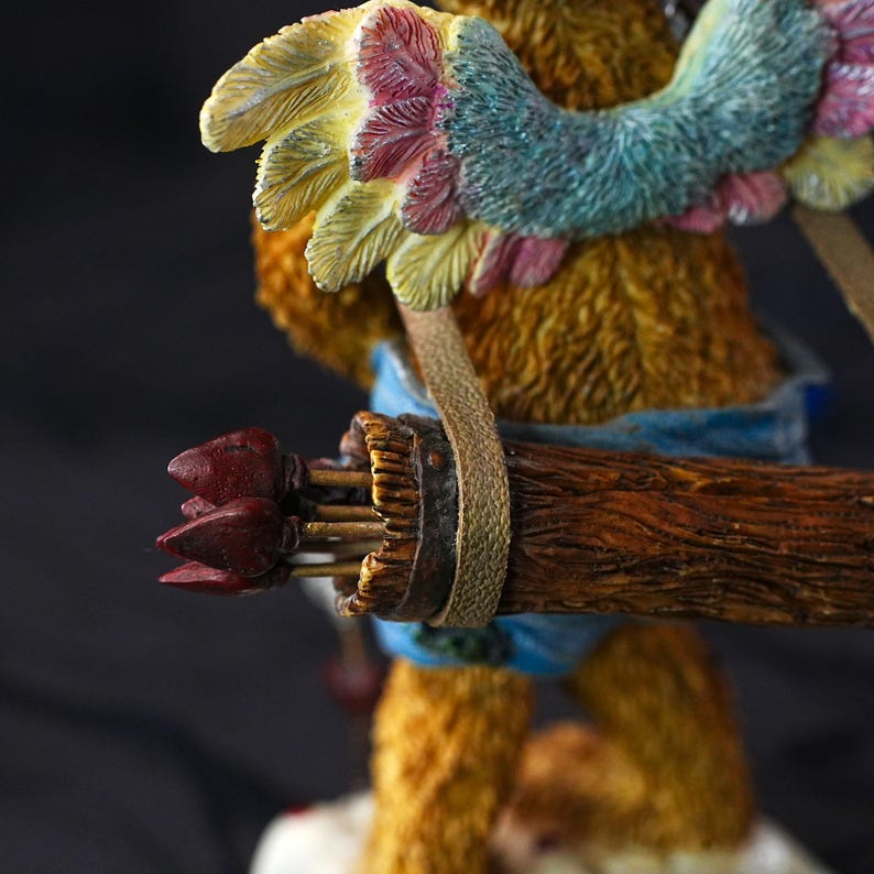 Puede incluir: Una figura de oso marr&oacute;n texturizado con grandes alas multicolores de plumas amarillas, rosas y azules. El oso lleva una prenda azul y porta un carcaj marr&oacute;n con textura de madera lleno de flechas con puntas de color rojo oscuro.