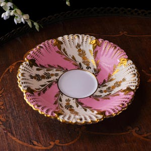 May include: A vintage pink and white porcelain saucer with gold trim and floral detailing. The saucer has a scalloped edge and a white center. The design features pink sections alternating with white sections, adorned with gold leaf patterns.