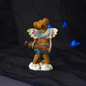 Puede incluir: Una figura de oso de peluche con textura marr&oacute;n de pie sobre una base de nube blanca. El oso presenta alas de colores pastel en tonos rosa, azul y amarillo, y lleva un carcaj de textura de madera marr&oacute;n lleno de flechas en la espalda.