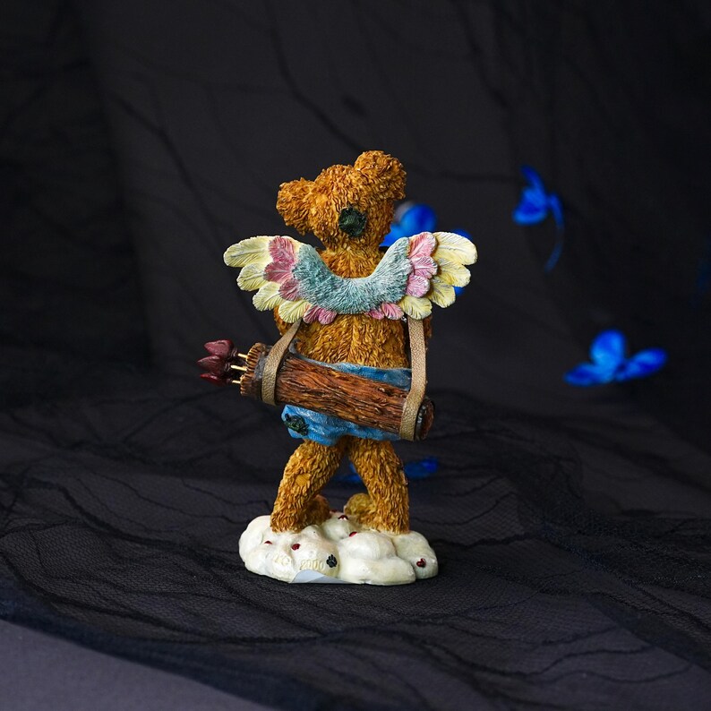 Puede incluir: Una figura de oso de peluche con textura marr&oacute;n de pie sobre una base de nube blanca. El oso presenta alas de colores pastel en tonos rosa, azul y amarillo, y lleva un carcaj de textura de madera marr&oacute;n lleno de flechas en la espalda.