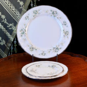 Pode incluir: Dois pratos de porcelana branca com um design floral e acabamento prateado. O prato maior é exibido em um suporte de metal, com um prato menor empilhado em cima. O padrão floral apresenta flores verdes claras e amarelas.