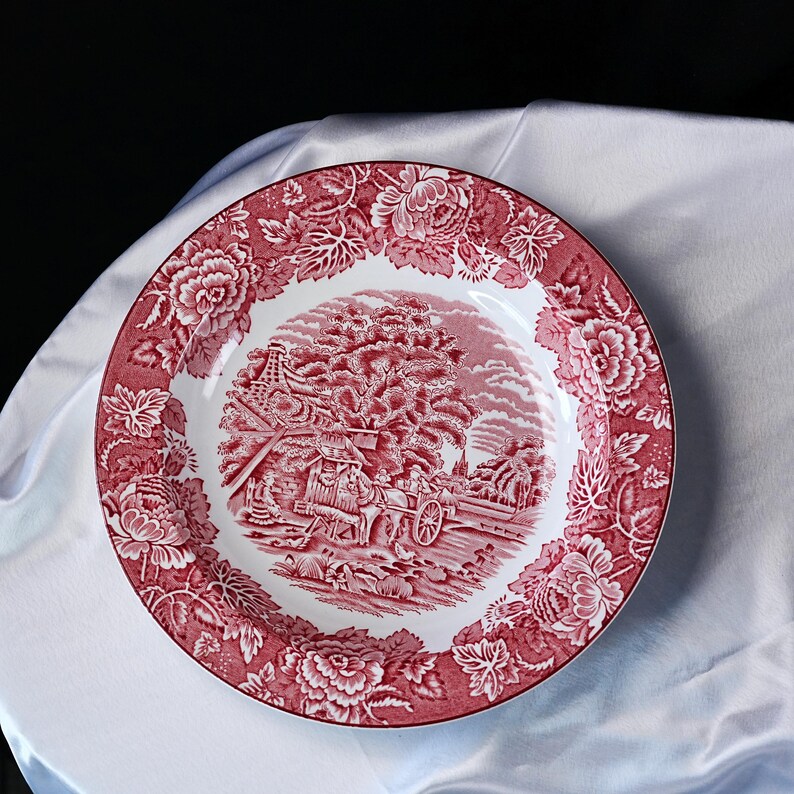 Op de afbeelding: Een ronde keramische bord met een donkerrood en wit transferpatroon. Het midden toont een landelijk tafereel met een paardenkoets, terwijl de brede rand is versierd met een dicht, herhalend bloemmotief van grote bloesems.