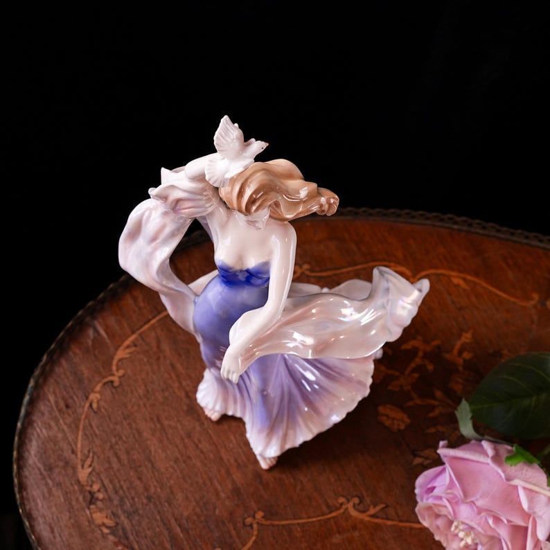 May include: A porcelain figurine of a woman in a flowing gown, with a dove perched on her shoulder. The dress transitions from a deep blue bodice to a soft lavender skirt. The figurine has light brown hair and is set against a dark background.