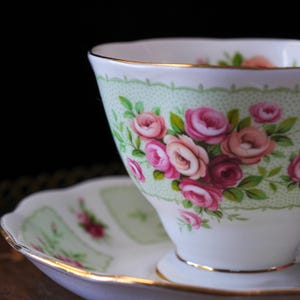 K&ouml;nnte beinhalten: Ein wei&szlig;es Tee-Set mit Tasse und Untertasse. Die Tasse hat einen hellgr&uuml;nen, gemusterten Rand und ist mit rosa und roten Rosen verziert. Beide Teile haben einen goldenen Rand.