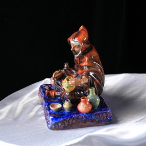 May include: A ceramic figurine of a robed figure crafting pottery, seated on a blue base. The figure wears a brown hooded robe and is surrounded by small, colourful vases and bowls. The base has a textured, glazed finish.