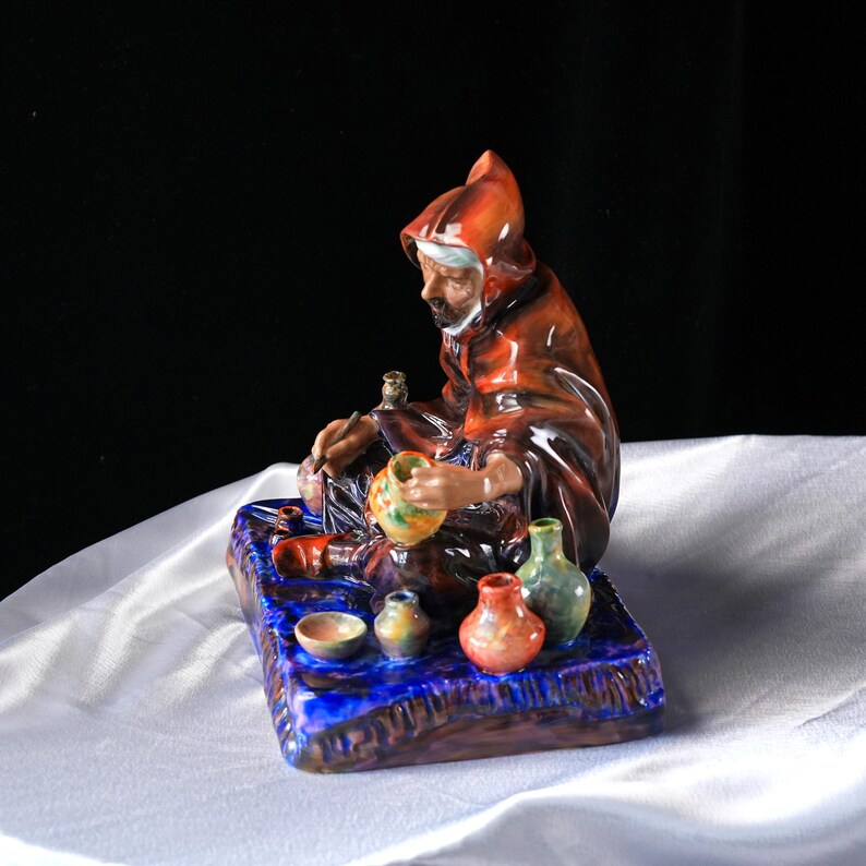 May include: A ceramic figurine of a robed figure crafting pottery, seated on a blue base. The figure wears a brown hooded robe and is surrounded by small, colourful vases and bowls. The base has a textured, glazed finish.