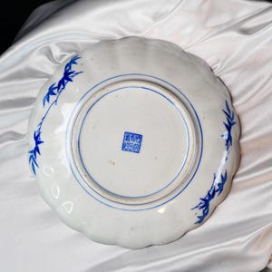 Puede incluir: Plato de cer&aacute;mica blanca con borde festoneado, con dise&ntilde;os de hojas de bamb&uacute; azules y un borde circular azul. El plato tiene un sello azul en el centro. Sobre una superficie de sat&eacute;n blanco.