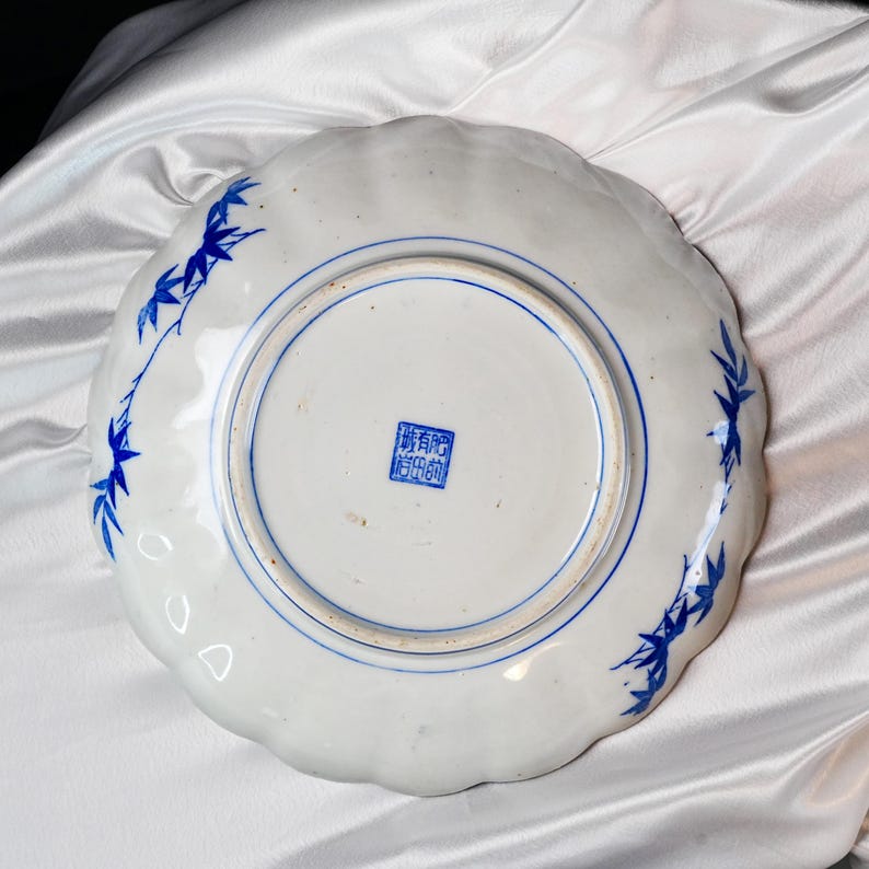 Puede incluir: Plato de cer&aacute;mica blanca con borde festoneado, con dise&ntilde;os de hojas de bamb&uacute; azules y un borde circular azul. El plato tiene un sello azul en el centro. Sobre una superficie de sat&eacute;n blanco.