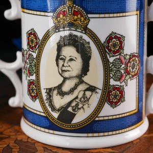 May include: A white ceramic mug featuring a central oval portrait of Queen Elizabeth The Queen Mother in black and white, topped with a gold crown. The design includes red and green floral motifs, a blue border, and gold trim accents.