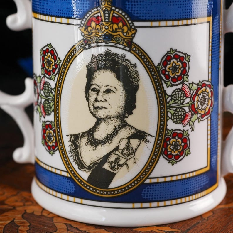 May include: A white ceramic mug featuring a central oval portrait of Queen Elizabeth The Queen Mother in black and white, topped with a gold crown. The design includes red and green floral motifs, a blue border, and gold trim accents.
