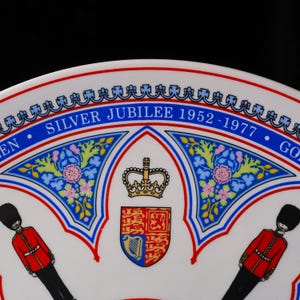 Puede incluir: Un plato conmemorativo de cer&aacute;mica blanca con el texto SILVER JUBILEE 1952-1977. El dise&ntilde;o incluye un escudo real central coronado, flanqueado por dos guardias con uniformes rojos y gorros de piel de oso negros, con detalles florales azules.