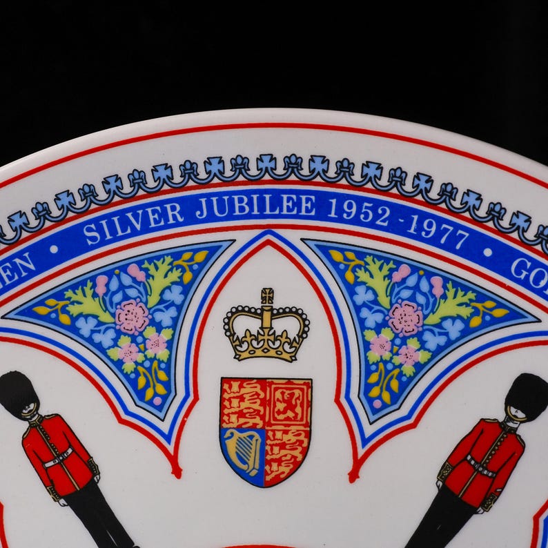 Puede incluir: Un plato conmemorativo de cer&aacute;mica blanca con el texto SILVER JUBILEE 1952-1977. El dise&ntilde;o incluye un escudo real central coronado, flanqueado por dos guardias con uniformes rojos y gorros de piel de oso negros, con detalles florales azules.