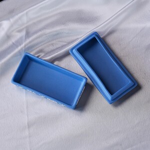 May include: Two open, rectangular blue ceramic trinket boxes. One box is upright, and the other is lying on its side. The boxes have a smooth, matte finish. The edges of the boxes are slightly raised. The boxes are on a white, textured surface.