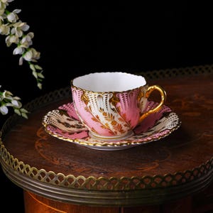 May include: A pink and gold teacup and saucer set. The teacup has a gold handle and rim, with a pink and white floral design. The saucer has a matching design. The set is displayed on a wooden table.