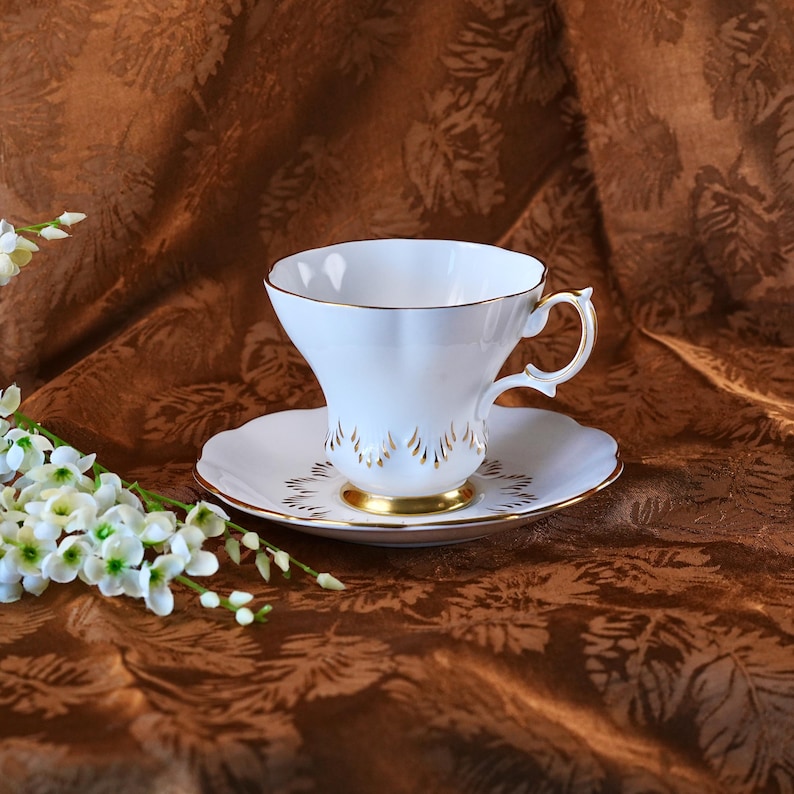 May include: A white teacup and saucer set with gold trim. The teacup has a flared shape and a delicate handle. The saucer features a matching gold design. The set is placed on a brown patterned fabric, with white flowers in the foreground.