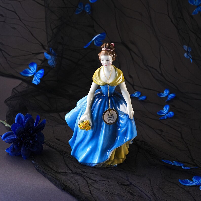 May include: A porcelain figurine of a person wearing a blue and yellow gown, holding a bouquet of yellow flowers. A gold-colored tag hangs from the neck, featuring the text Royal Doulton, Bone China, and Hand Decorated, England.
